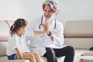 Gives high five to the little girl. Senior male doctor with grey hair and beard in white coat is indoors in clinic photo