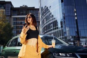 With smartphone. Young fashionable woman in burgundy colored coat at daytime with her car photo
