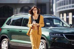 con bebida en las manos. joven mujer de moda con abrigo de color burdeos durante el día con su coche foto