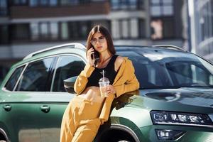 With smartphone. Young fashionable woman in burgundy colored coat at daytime with her car photo