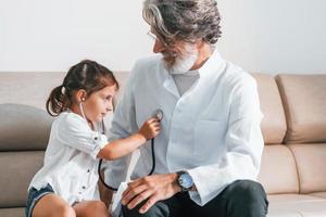 Little girl playing with stethoscope. Senior male doctor with grey hair and beard in white coat is indoors in clinic photo
