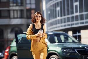 Against business building. Young fashionable woman in burgundy colored coat at daytime with her car photo