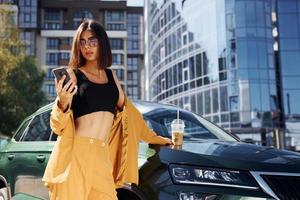 With smartphone. Young fashionable woman in burgundy colored coat at daytime with her car photo