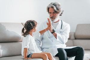 Gives high five to the little girl. Senior male doctor with grey hair and beard in white coat is indoors in clinic photo