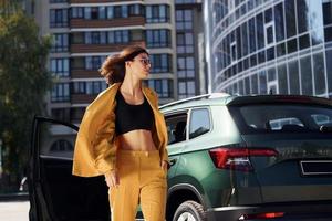 camina hacia adelante desde el automóvil. joven mujer de moda con abrigo de color burdeos durante el día con su coche foto