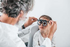 Senior male ophthalmologist with grey hair and beard in white coat is indoors in clinic testing sight of little girl photo