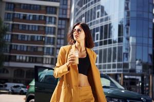 Against business building. Young fashionable woman in burgundy colored coat at daytime with her car photo