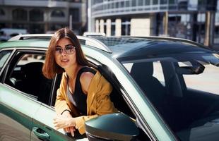 Looks out from automobile. Young fashionable woman in burgundy colored coat at daytime with her car photo