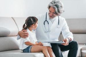 Talks with little girl. Working with customer. Senior male doctor with grey hair and beard in white coat is indoors in clinic photo