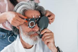 Getting tested by woman worker. Senior man with grey hair and beard is in ophthalmology clinic photo