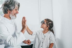 Senior male ophthalmologist with grey hair and beard in white coat is indoors in clinic testing sight of little girl photo