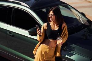 Takes break with cup of coffee. Young fashionable woman in burgundy colored coat at daytime with her car photo