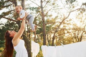 Standing near white clothes that hanging on the rope to dry. Young mother with her little son is outdoors in the forest. Beautiful sunshine photo