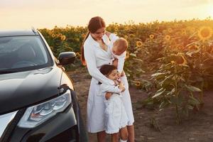 cerca de un coche negro moderno. la joven madre con su pequeño hijo y su hija está al aire libre en el campo agrícola. hermoso amanecer foto