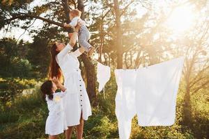 ropa blanca colgada de la cuerda para secarse. la joven madre con su pequeña hija y su hijo está al aire libre en el bosque. hermoso amanecer foto