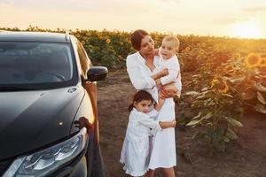 Near modern black car. Young mother with her little son and daughter is outdoors in the agricultural field. Beautiful sunshine photo