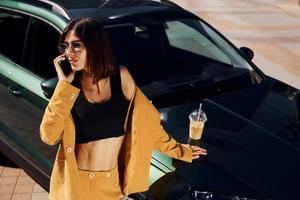 With smartphone. Young fashionable woman in burgundy colored coat at daytime with her car photo