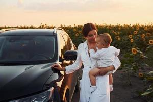 cerca de un coche negro moderno. la joven madre con su hijo pequeño está al aire libre en el campo agrícola. hermoso amanecer foto
