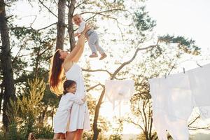 ropa blanca colgada de la cuerda para secarse. la joven madre con su pequeña hija y su hijo está al aire libre en el bosque. hermoso amanecer foto