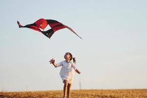 Happy little girl running with red kite outdoors on the field at summertime photo