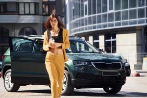 Against business building. Young fashionable woman in burgundy colored coat at daytime with her car photo