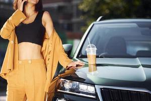 Takes break with cup of coffee. Young fashionable woman in burgundy colored coat at daytime with her car photo