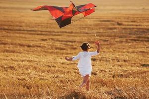 Happy little girl running with red kite outdoors on the field at summertime photo