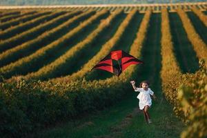 Active little girl in white clothes running with red kite on the field photo