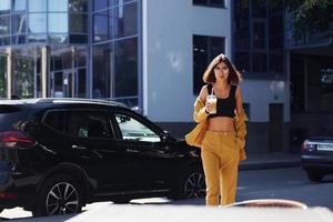 Coffee break. Young fashionable woman in burgundy colored coat at daytime with her car photo