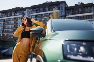 With smartphone. Young fashionable woman in burgundy colored coat at daytime with her car photo