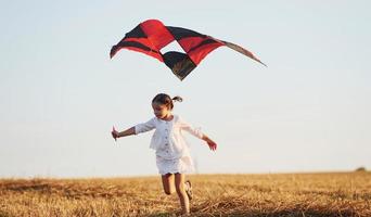 Happy little girl running with red kite outdoors on the field at summertime photo