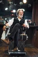 se sienta en la silla de la barbería. un anciano moderno y elegante con pelo gris y barba está en el interior foto