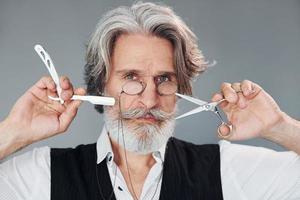 Barbershop tools. Against grey background. Stylish modern senior man with gray hair and beard is indoors photo