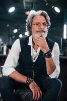 Sits on the chair in barbershop. Stylish modern senior man with gray hair and beard is indoors photo