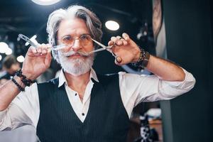 tiene equipo de barbería. un anciano moderno y elegante con pelo gris y barba está en el interior foto