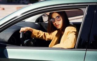 Happy automobile owner sits inside of vehicle. Young fashionable woman in burgundy colored coat at daytime with her car photo