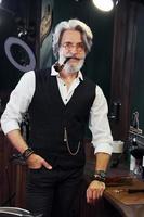 de pie y fumando en la barbería. un anciano moderno y elegante con cabello gris y barba está en el interior foto