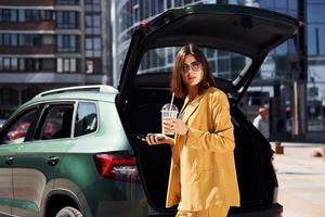 café y teléfono en las manos. joven mujer de moda con abrigo de color burdeos durante el día con su coche foto