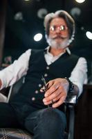 Sits on the chair in barbershop. Stylish modern senior man with gray hair and beard is indoors photo