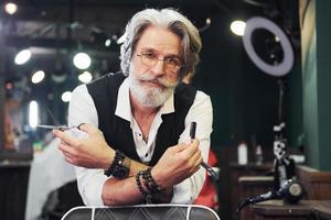 Holds barbershop equipment. Stylish modern senior man with gray hair and beard is indoors photo