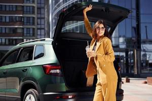 Holding drink and smiling. Young fashionable woman in burgundy colored coat at daytime with her car photo