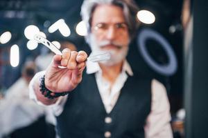 Holds barbershop equipment. Stylish modern senior man with gray hair and beard is indoors photo