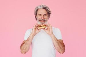 Eats delicious burger. Stylish modern senior man with gray hair and beard is indoors photo