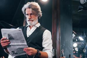 leyendo el periódico. un anciano moderno y elegante con pelo gris y barba está en el interior foto
