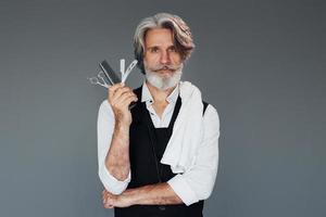 Professional barbershop worker. Against grey background. Stylish modern senior man with gray hair and beard is indoors photo