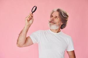 Using magnifying glass. Stylish modern senior man with gray hair and beard is indoors photo