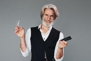 Professional barbershop worker. Against grey background. Stylish modern senior man with gray hair and beard is indoors photo