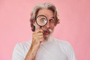 Using magnifying glass. Stylish modern senior man with gray hair and beard is indoors photo