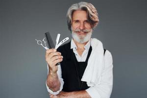 trabajador de barbería profesional. contra un fondo gris. un anciano moderno y elegante con cabello gris y barba está en el interior foto