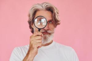 Using magnifying glass. Stylish modern senior man with gray hair and beard is indoors photo